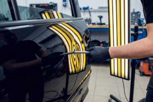 Technician examines a dented car door under bright reflection lighting beside precision repair tools.