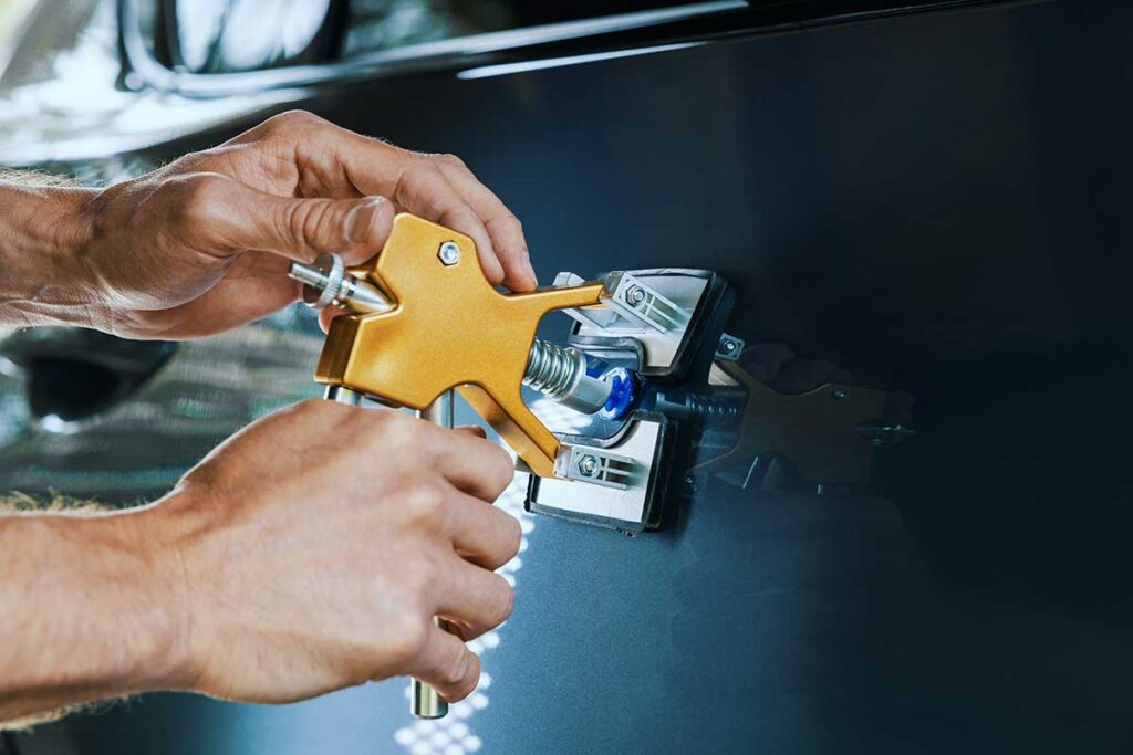 Technician uses a slim metal tool behind a car door while inspecting the panel under bright reflection lighting.